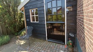 A dark wooden house with a black door and windows, a small patio with a metal chair, and a brick wall. - Romantic Cottage; hier kom je tot rust! (Ulft)