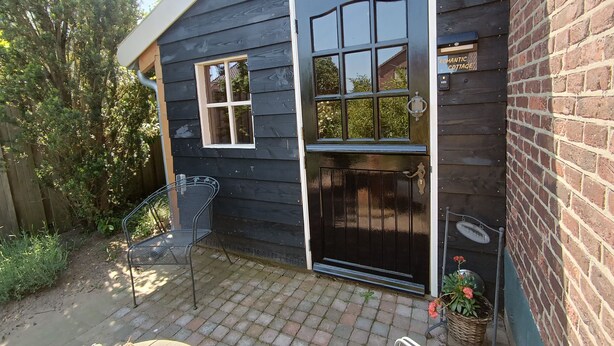 Ein dunkles Holzhaus mit einer schwarzen Tür und Fenstern, einer kleinen Terrasse mit einem Metallstuhl und einer Ziegelwand.