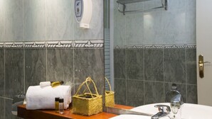 Baño con pared de mármol, dispensador de toallas, secador de manos, tendedero para toallas y encimera de madera con artículos de aseo.