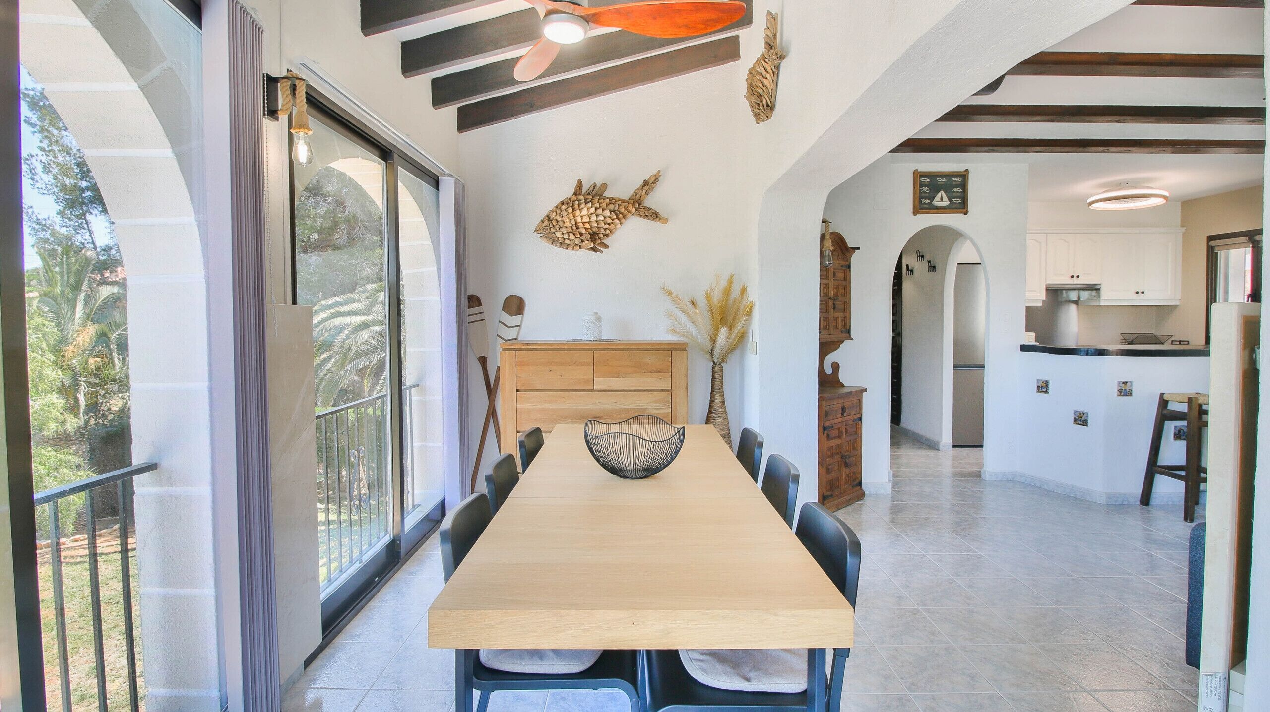 A dining area with a long wooden table, chairs, a decorative fish wall art, and a view through a glass door to a balcony.