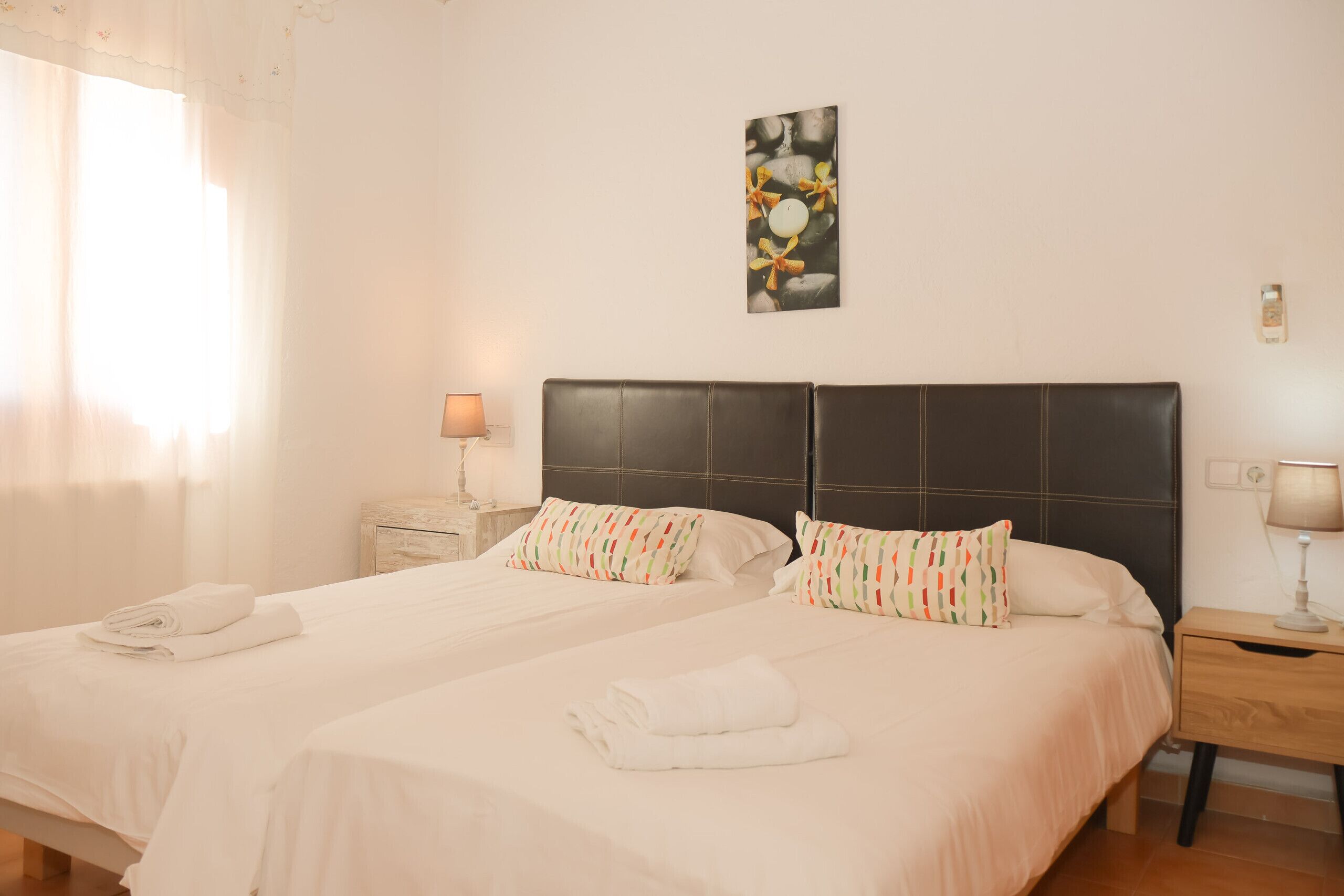 Une chambre d’hôtel avec deux lits, chacun équipé d’un oreiller et d’une serviette roulée. On y trouve une table de chevet avec une lampe, une œuvre d’art accrochée au mur et une fenêtre avec des rideaux.