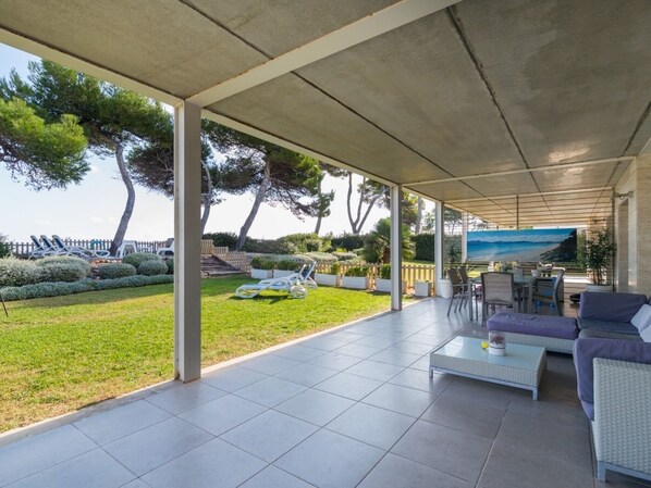 Un patio couvert avec vue sur la mer, doté de mobilier d’extérieur et bordé d’un jardin aménagé.