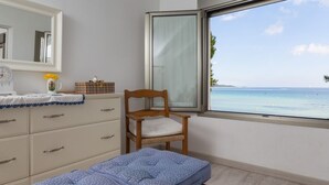 A bedroom with a wooden chair, a bed with a blue polka dot cover, a dresser with drawers, a mirror, and a window with a view of the sea. - Las Gaviotas - Spectacular beachfront villa with garden in Platja de Muro (Muro (Platja de Muro))