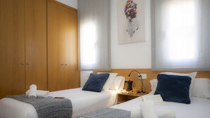 A hotel room with two beds, a wooden wardrobe, a wall-mounted artwork, and a bedside table with a lamp and a coffee maker.