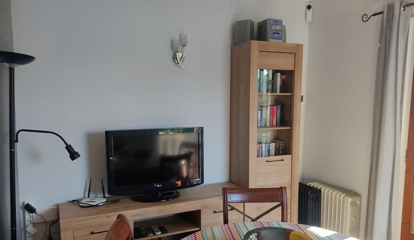 Um quarto com uma TV sobre um suporte de madeira, uma estante, uma cadeira e um radiador.
