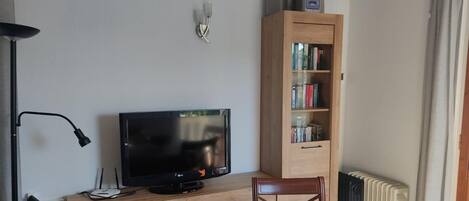 Um quarto com uma TV sobre um suporte de madeira, uma estante, uma cadeira e um radiador.