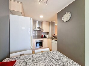 A compact kitchen with a stove, oven, and sink, a clock on the wall, and a patterned bedspread.