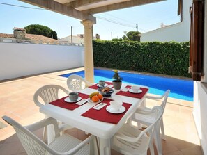 Un coin repas avec une table dressée pour deux personnes, une piscine en arrière-plan et une haie le long du mur.