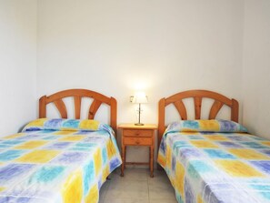 Une chambre avec deux lits en bois, dotés de draps aux couleurs vives, une petite table de chevet en bois avec une lampe, et un sol carrelé.
