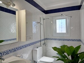 Une salle de bain avec des toilettes blanches, un lavabo, une douche et une fenêtre.