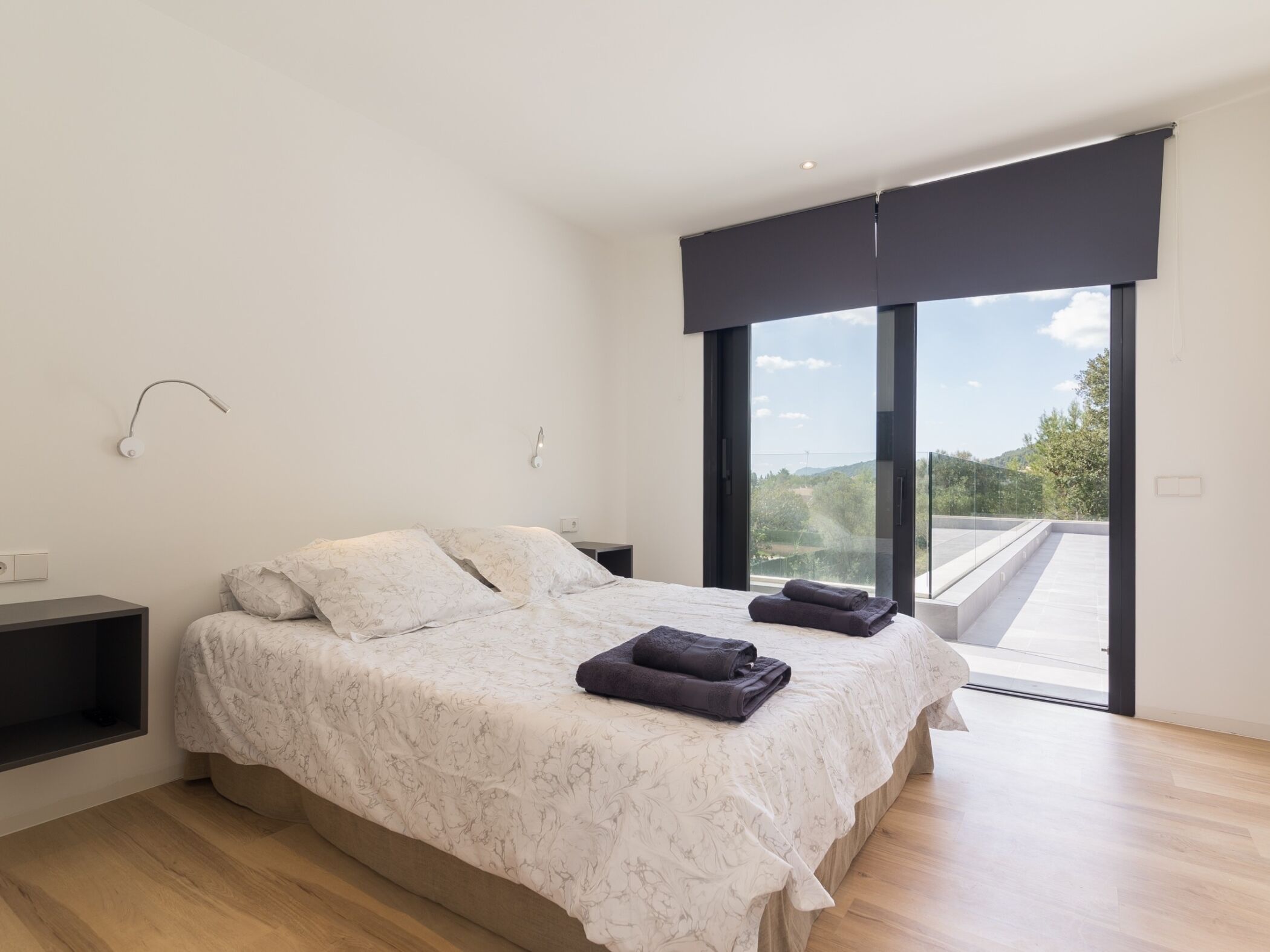 Ein modernes Schlafzimmer mit einem großen Bett, einem Nachttisch, einer Wandlampe und Blick ins Freie durch eine Schiebetür.