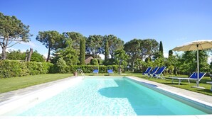Ein rechteckiger Swimmingpool mit klarem, blauem Wasser, umgeben von einem gepflegten Rasen und Bäumen. Auf der rechten Seite befinden sich mehrere Liegestühle und ein Sonnenschirm.