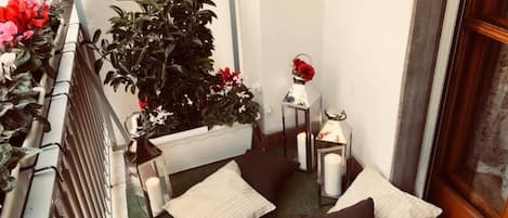 Ein kleiner Balkon mit einer grünen Matte, zwei weißen und einem schwarzen Kissen, einer Topfpflanze und einem Laternenlicht mit einer roten Rose.
