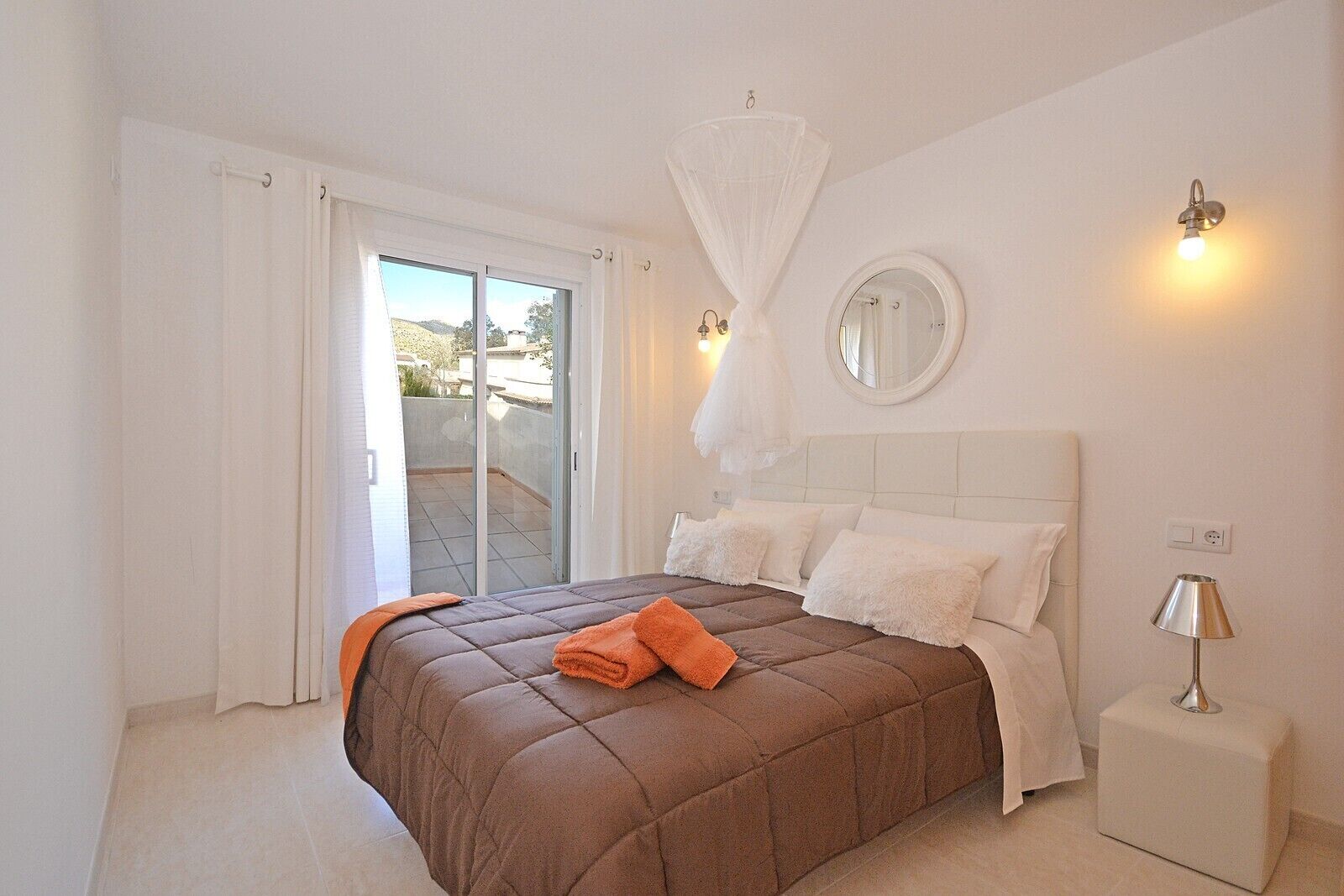 A bedroom with a large bed, white curtains, a balcony, a round mirror, and a nightstand with a lamp.