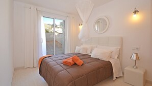Un dormitorio con una cama grande, cortinas blancas, un balcón, un espejo redondo y una mesita de noche con lámpara.