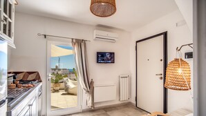 A modern hotel room with a kitchen, a TV mounted on the wall, a bed with a wooden headboard, and a view of the city through the window. - Il Nido - Le Case di Valentina (Gallipoli)