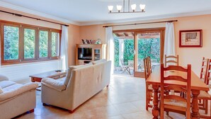 Amplia sala de estar con sofá, mesa de comedor con sillas, televisor y vistas al exterior a través de ventanas grandes.