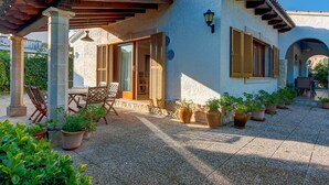 Un patio techado con mobiliario exterior, plantas en macetas y vista al interior del edificio.