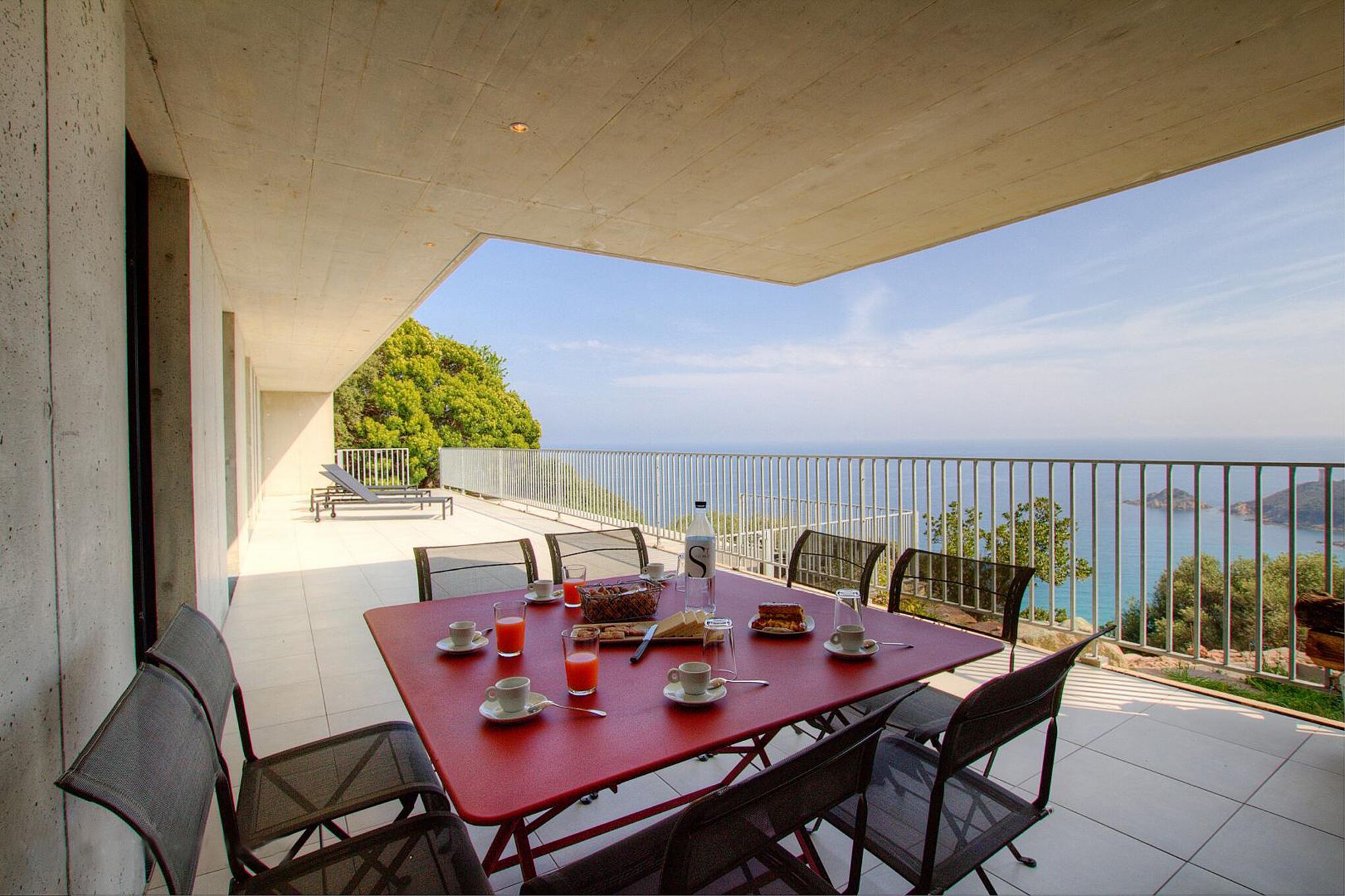 A red table set with breakfast items and a scenic view of the sea.
