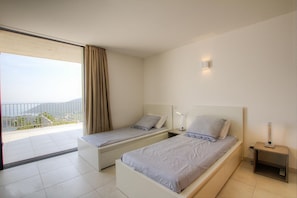 A modern bedroom with two single beds, a nightstand, and a bedside table with a lamp. A large window with curtains offers a view of the outdoors. - VILLA ARCHITECTE 5☆ 180° SEA VIEW and TOUR GÉNOISE. HEATED POOL. INTIMACY. (Conca)