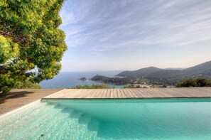 On peut admirer une piscine aux eaux cristallines, bordée d’une terrasse en bois, entourée d’une végétation luxuriante et offrant une vue imprenable sur un paysage côtier avec des collines et la mer.
