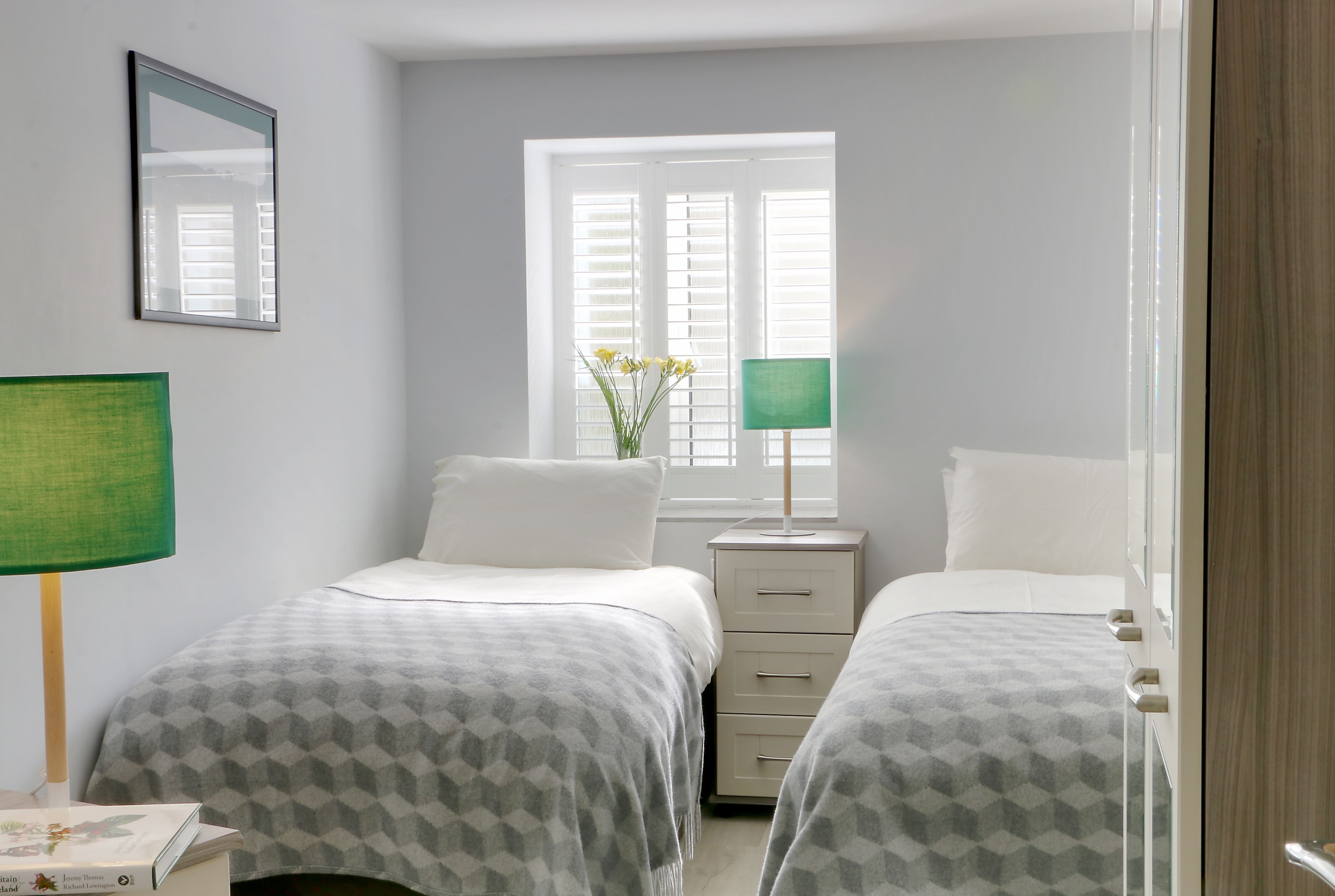 A bedroom with two beds, a nightstand, a lamp, a mirror, and a window with blinds.