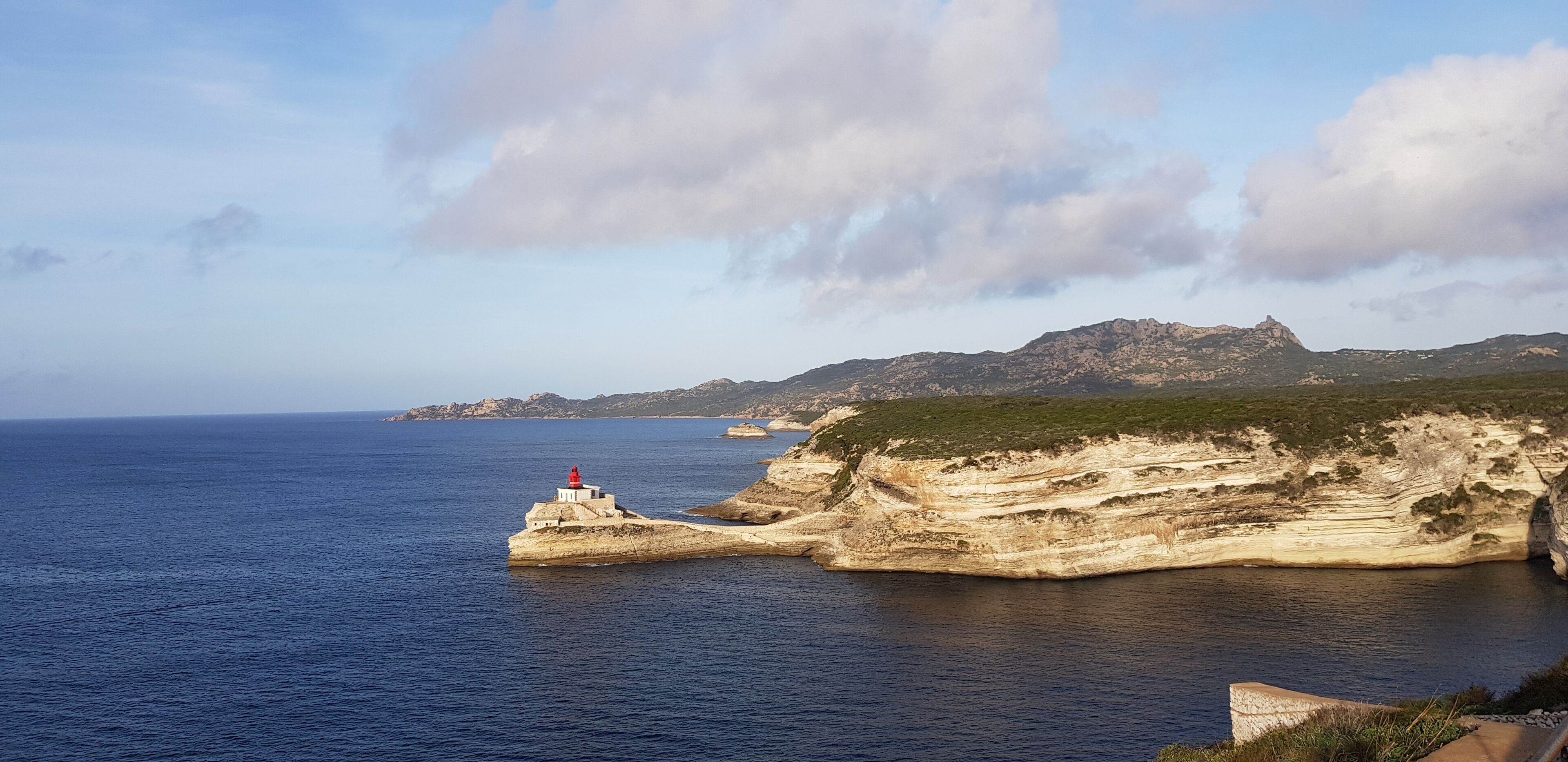 A coastal landscape with a lighthouse on a rocky outcrop, clear skies, and calm sea.