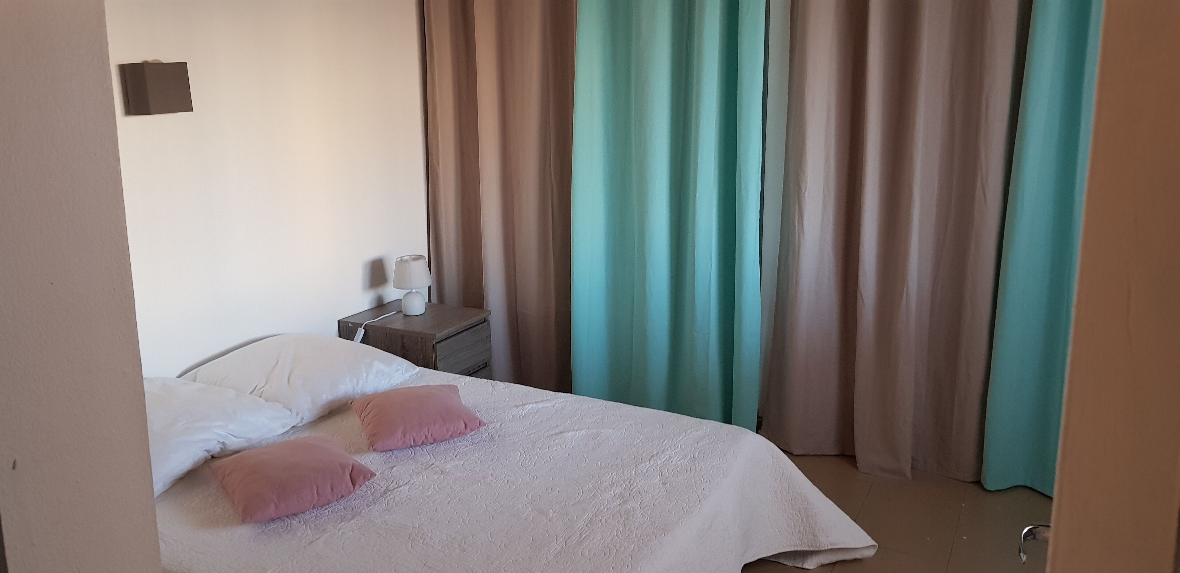 A neatly made bed with white and pink pillows, a bedside table with a lamp, and curtains in teal and beige colors.