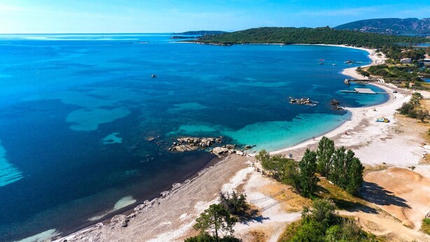 Vista aérea de una zona costera con aguas azules cristalinas, una playa arenosa y un paisaje montañoso al fondo.