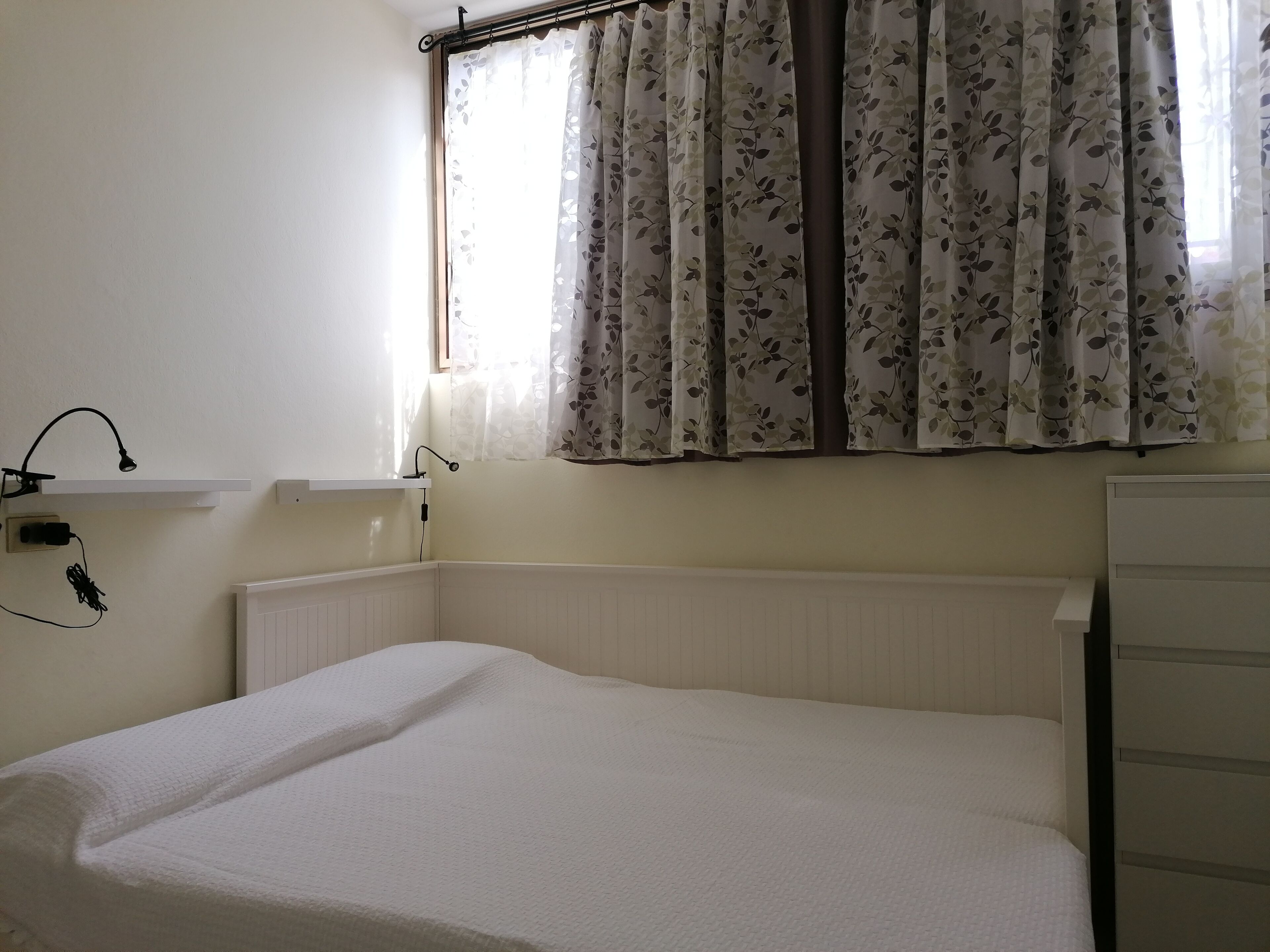 Una cama pequeña y bien hecha en una habitación con un radiador, una ventana con cortinas florales y un estante en la pared con una lámpara y toma de corriente.