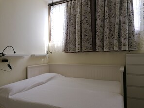 Una cama pequeña y bien hecha en una habitación con un radiador, una ventana con cortinas florales y un estante en la pared con una lámpara y toma de corriente.
