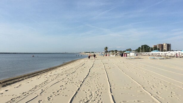 Una extensa playa arenosa con huellas que conducen al agua, algunas personas caminando y edificios al fondo.