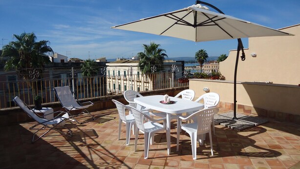 A terrace with a table and chairs, a large umbrella, and a view of the sea and buildings. - Ortigia, comfortable Baron house (Siracusa)