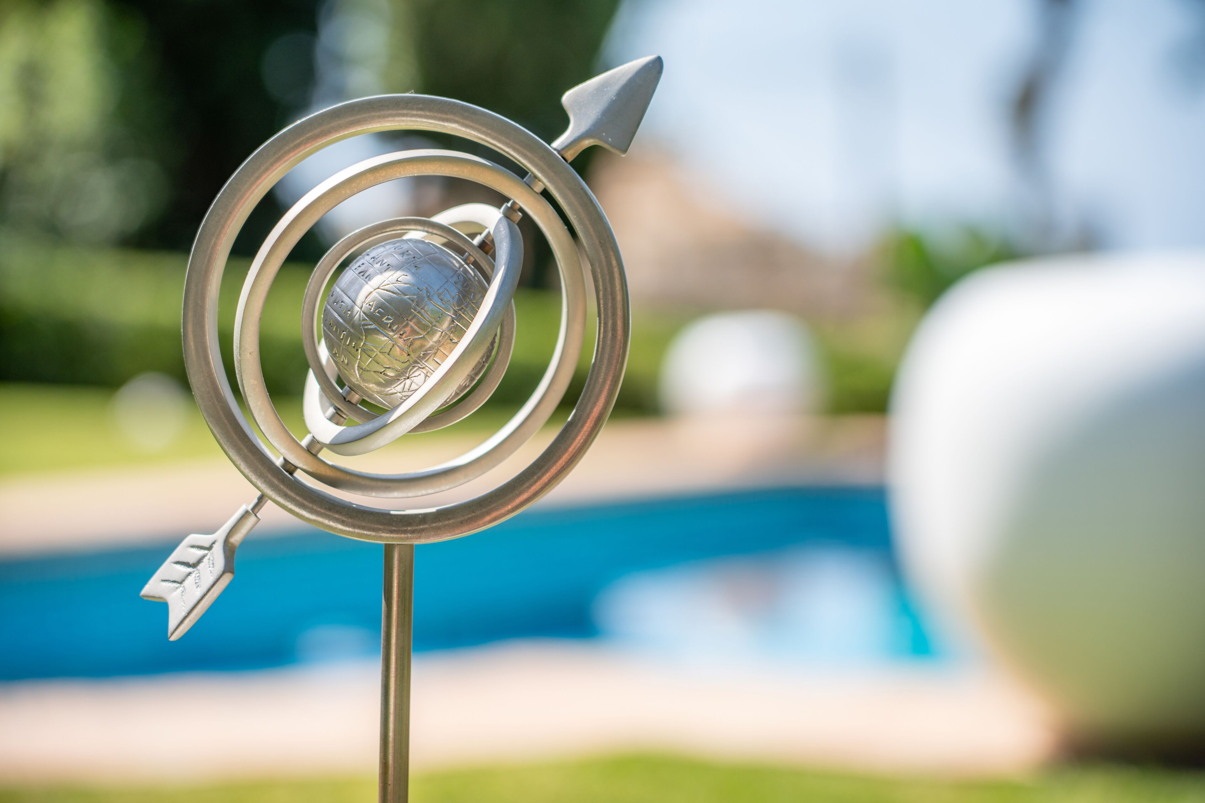 A metallic decorative object with a spherical center, possibly a sculpture, set against a blurred background featuring a swimming pool.
