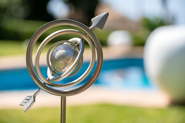 A decorative metal sculpture with a globe at its center, set against a blurred background of a swimming pool and greenery.