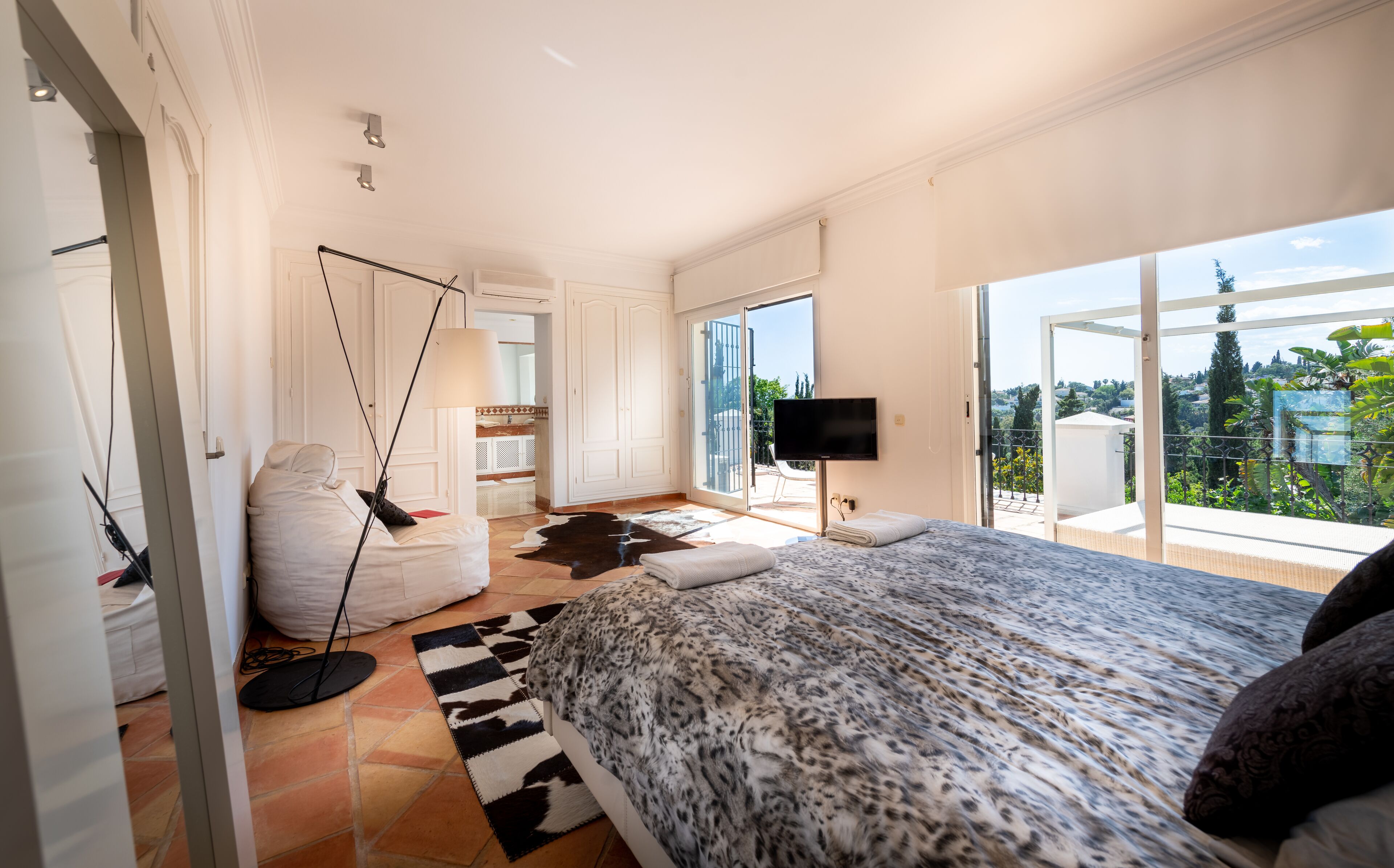 A spacious bedroom with a large bed, a TV, and a balcony with a view of greenery.