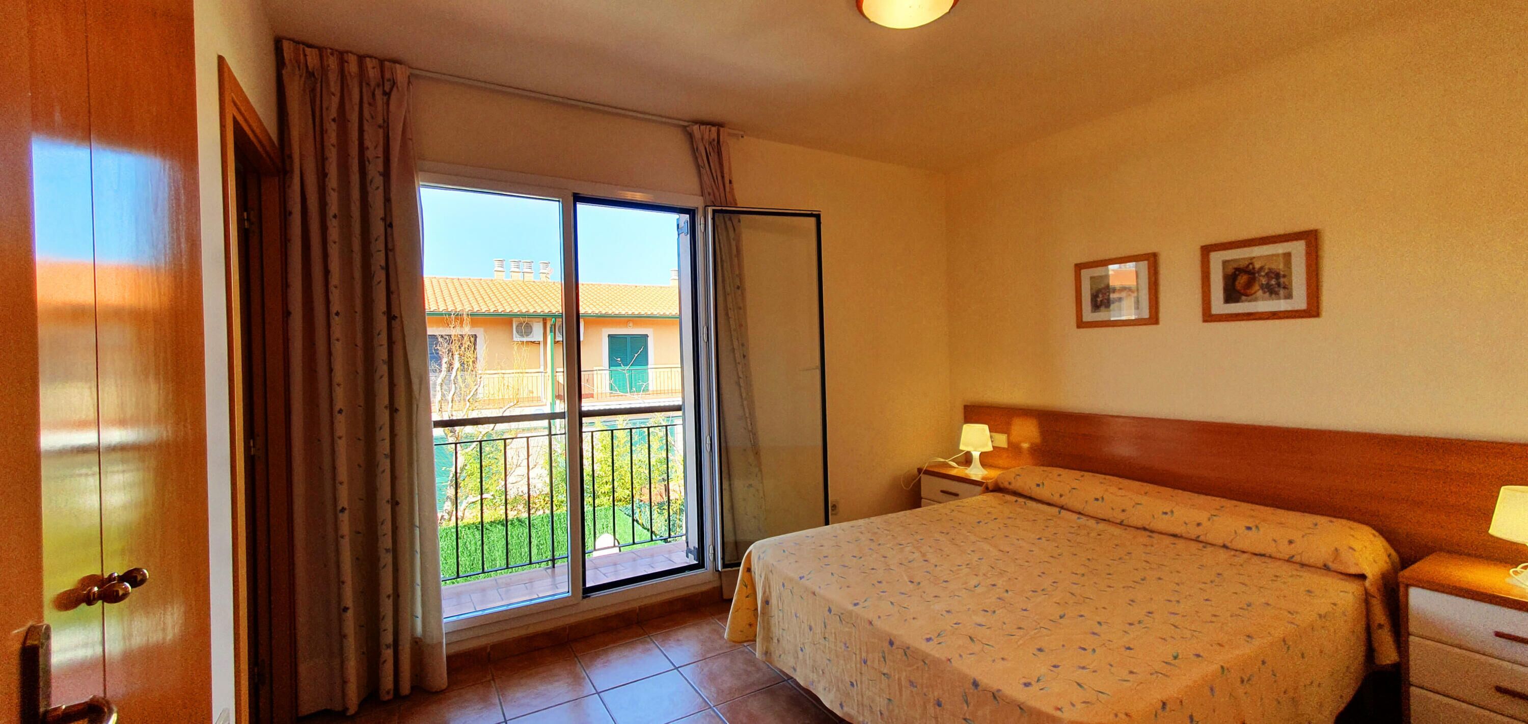 Habitación de hotel con cama, mesitas de noche, ventana con cortinas y vistas a un edificio y zonas verdes.