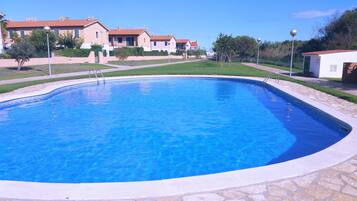 A swimming pool with a stone border, surrounded by a well-maintained lawn and a row of houses in the background.