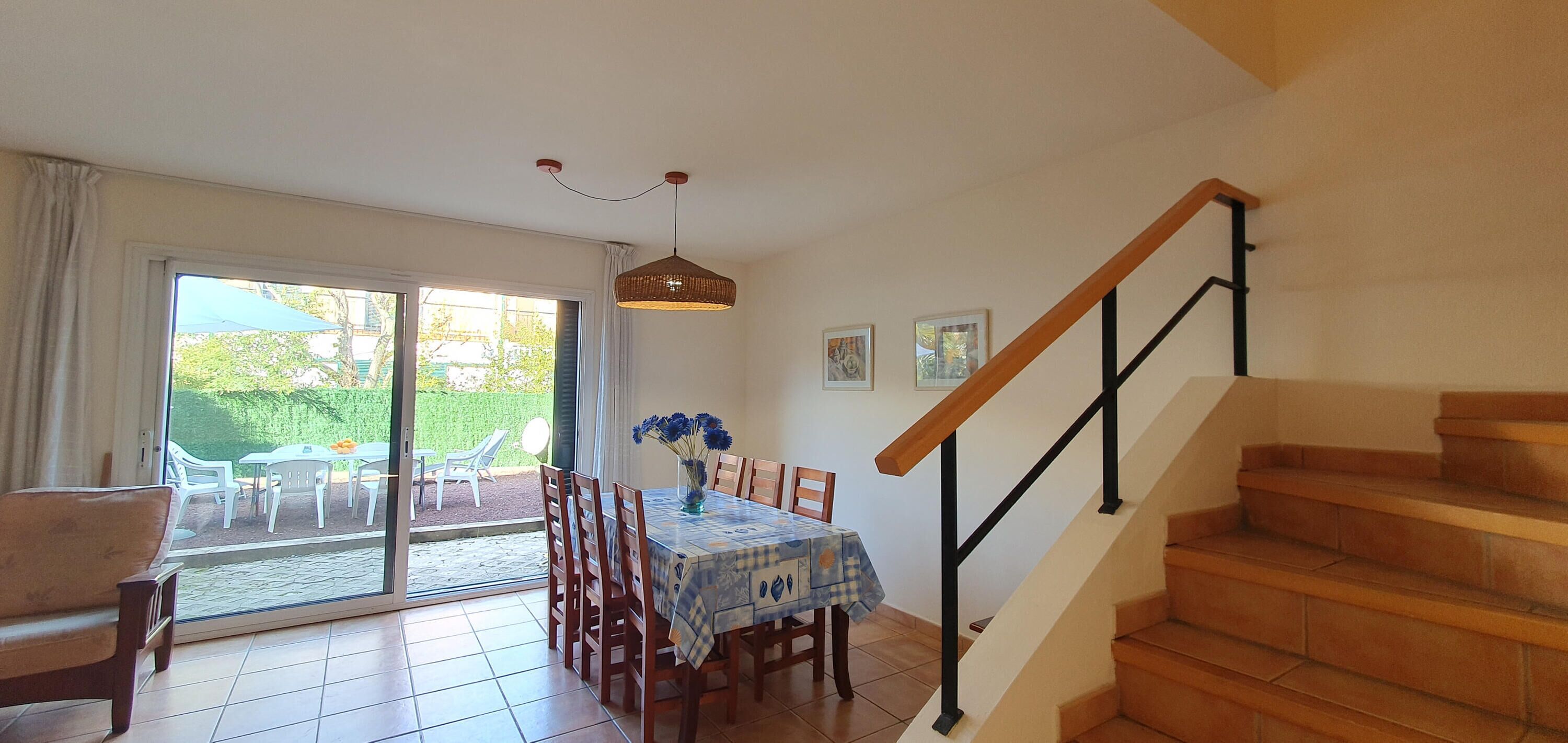 Un comedor con una mesa dispuesta para cuatro personas, una escalera de madera y vistas al jardín a través de la puerta de vidrio.