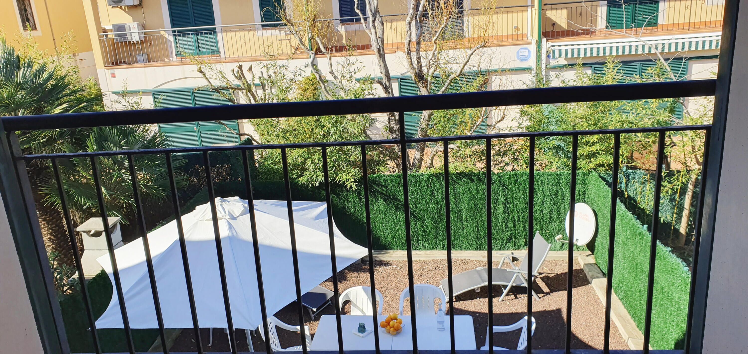 Un balcón con mesa y sillas blancas, un parasol blanco y un pequeño jardín con vegetación y seto.