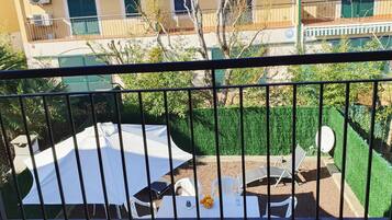 Un balcón con mesa y sillas blancas, un parasol blanco y un pequeño jardín con vegetación y seto.