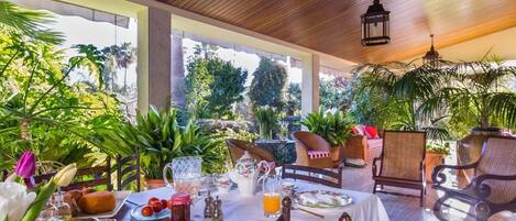 Un espace extérieur abrité, aménagé pour les repas, avec des tables et des chaises en bois, entouré d’une végétation luxuriante et de plantes.