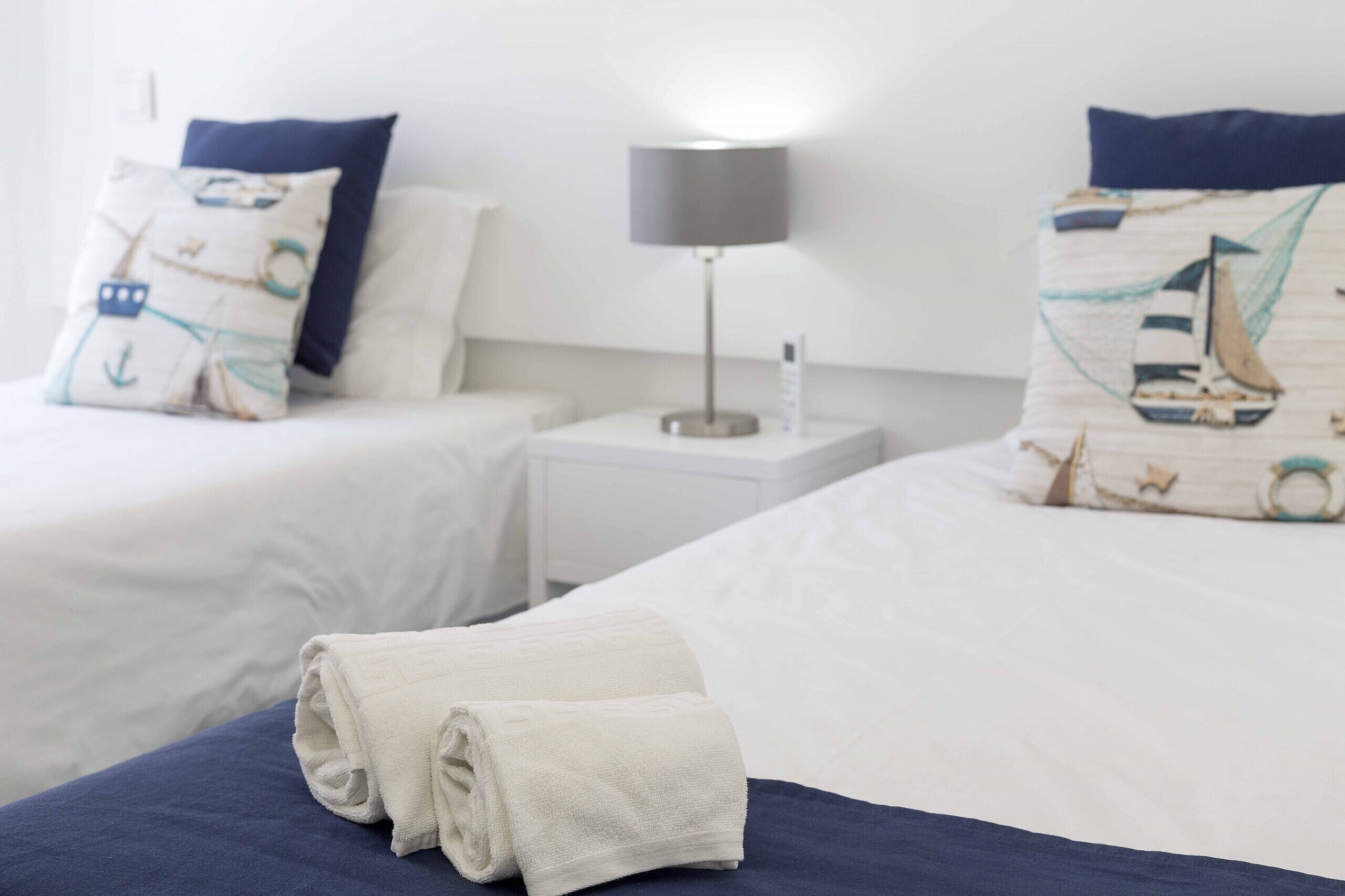 A hotel room with two beds, a nightstand with a lamp, and a folded towel on the bed.