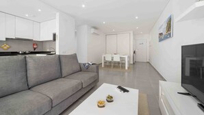 Een moderne woonkamer met een grijze bank, een bijzettafeltje met schalen, een televisie en een eethoek op de achtergrond.