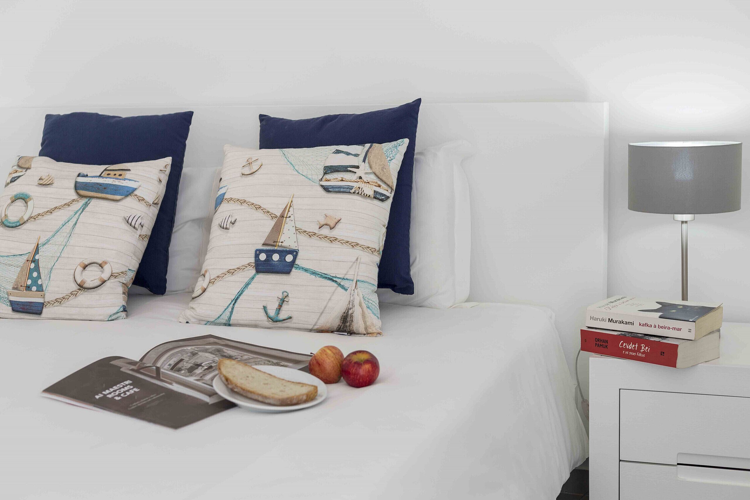 A neatly made bed with two pillows, a bedside table with a lamp, a book, and two apples.