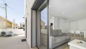 Een moderne woonkamer met een grijze bank, een witte salontafel en een glazen deur die uitkomt op een balkon met ligbedden.