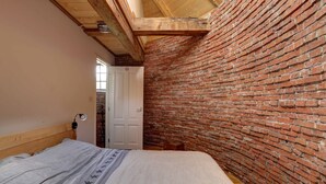 Ein Schlafzimmer mit Backsteinwand, Holzbalkendecke, einem Bett mit grauer Decke und einer weißen Tür.
