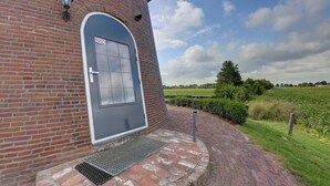 Ein Backsteinhaus mit grauer Tür, einer kleinen Veranda und einem grünen Feld im Hintergrund.