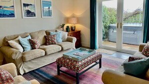 Un salon avec un canapé beige, un pouf à motifs et une table basse. On peut voir des tableaux encadrés au mur, une lampe sur une table d’appoint, et une vue sur un balcon agrémenté de plantes.