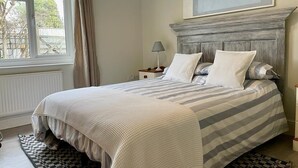 Une chambre à coucher avec un lit, une table de chevet, une lampe, une fenêtre avec des rideaux et un tableau au mur.